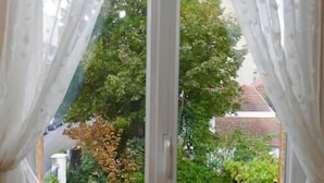 A window with white curtains, a view of a balcony with a metal railing, and greenery outside. - Chalet de Paris (Fontenay-sous-Bois)