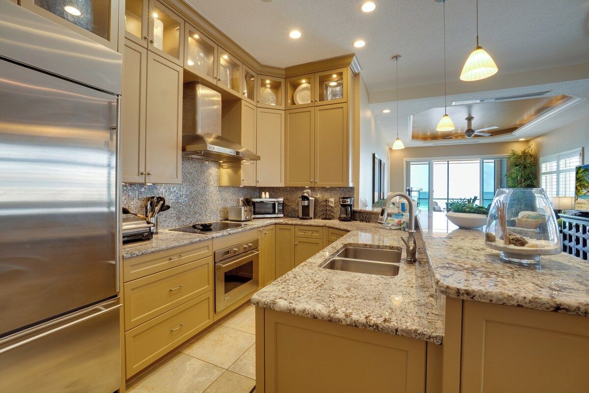 A modern kitchen with stainless steel appliances, beige cabinetry, granite countertops, and pendant lighting.
