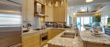 A modern kitchen with stainless steel appliances, beige cabinetry, granite countertops, and pendant lighting.