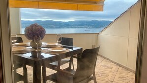 Una mesa de comedor dispuesta con platos, vasos y un florero en un balcón con vista a un lago y montañas.