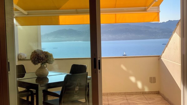 A balcony with a table and chairs, a vase with flowers, and a view of the sea and mountains.