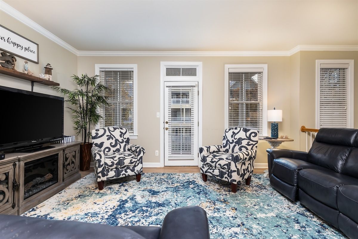 Un salon comprenant un téléviseur, deux fauteuils à motifs, un canapé en cuir noir, un tapis à motifs bleus et blancs, et une porte avec des volets blancs.