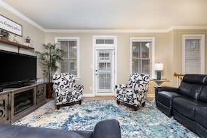 A living room with a television, two patterned armchairs, a black leather sofa, a blue and white patterned rug, and a door with white shutters. - Sunset Island 6 Island Edge Drive (Ocean City)