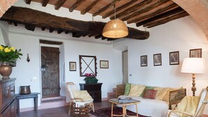 A rustic interior with a wooden ceiling, a woven light fixture, a sofa, two wicker chairs, a small table, and a patterned rug.