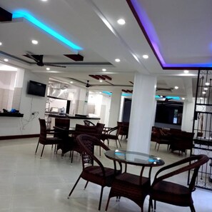 A modern interior with a dining area featuring round tables and wicker chairs, a kitchen area with a TV, and a ceiling with blue LED lighting. - CaliTwapa Hotel & Restaurant (Mtwapa)