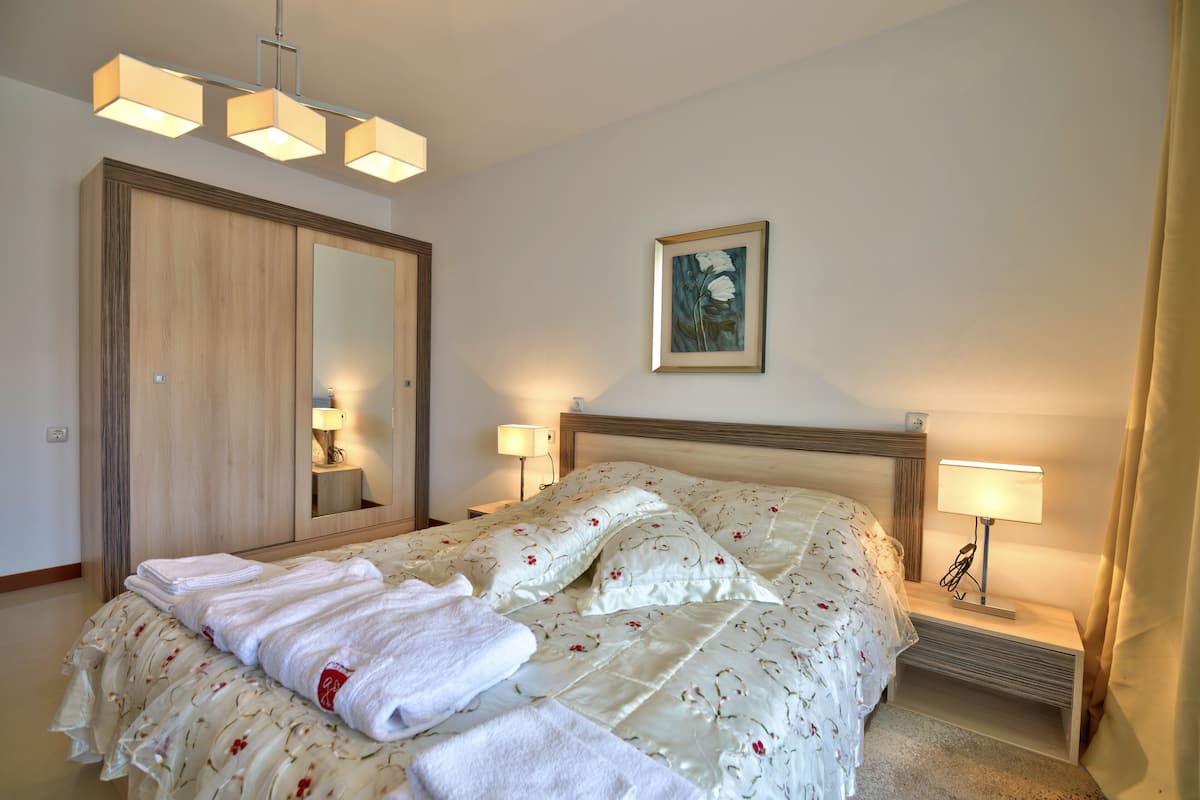 A neatly made bed with a floral duvet, a wooden wardrobe, a bedside table with a lamp, and a wall-mounted painting.