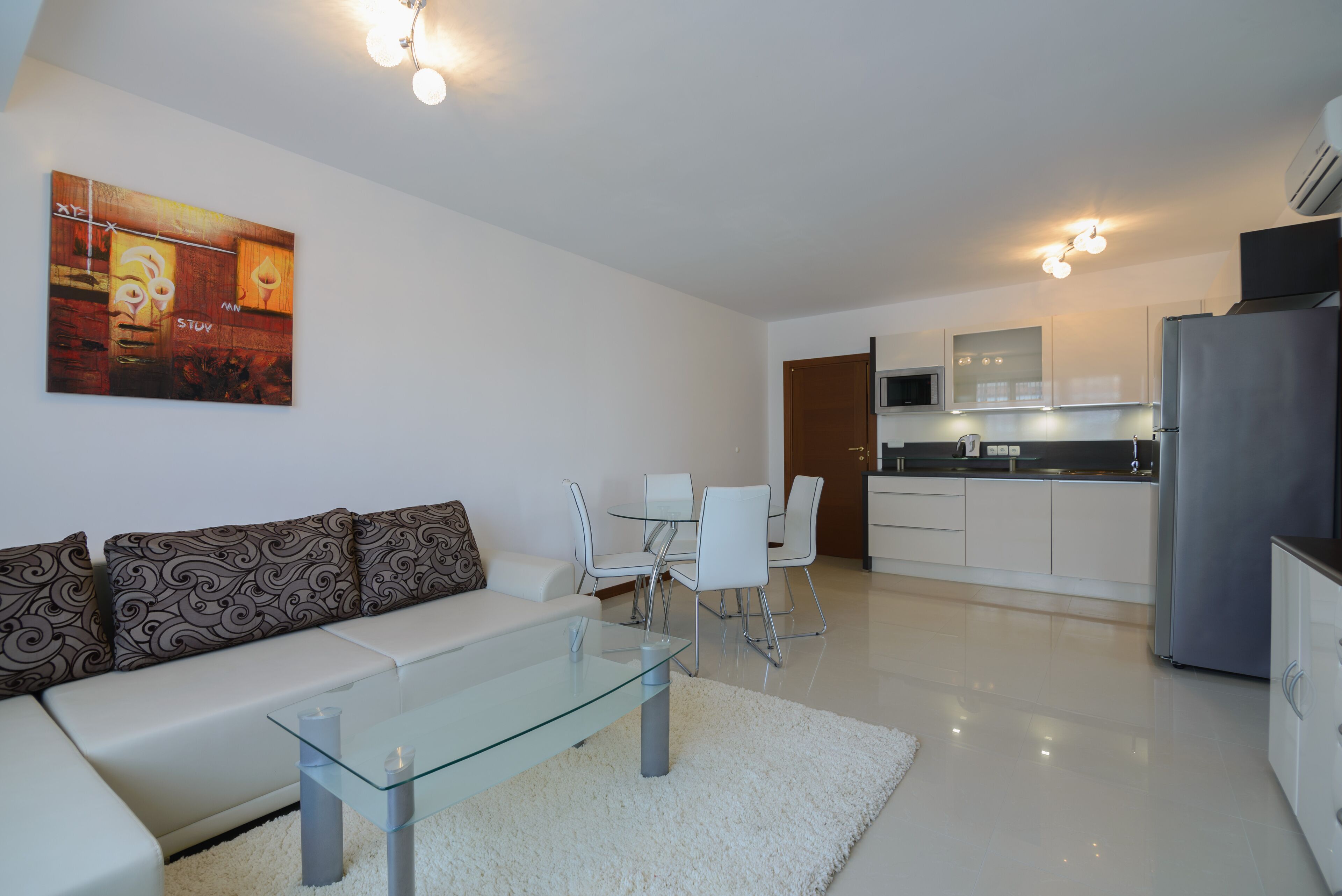 Uma sala de estar moderna com uma mesa de centro de vidro, sofá branco e almofadas estampadas. Uma área de jantar com cadeiras brancas e uma cozinha com eletrodomésticos de aço inoxidável e armários brancos.