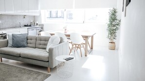 A modern kitchen with white cabinets, a dining area with a wooden table and chairs, and a grey sofa with a remote control on a side table.