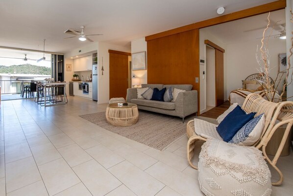 A spacious living area with a sofa, coffee table, and wicker chairs, a kitchen with a dining area, and a balcony with a view. - 1 Bright Point Apartment 1307 (Nelly Bay)