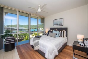 A bedroom with a large bed, a ceiling fan, a view of a body of water, and a balcony with plants. - 1 Bright Point Apartment 1307 (Nelly Bay)
