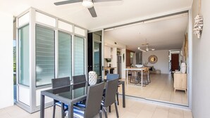 A modern dining area with a glass table and chairs, a ceiling fan, and a view into the kitchen.
