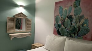Un dormitorio con una cama, una mesita de noche, un espejo con contraventanas y una pintura de cactus en la pared.