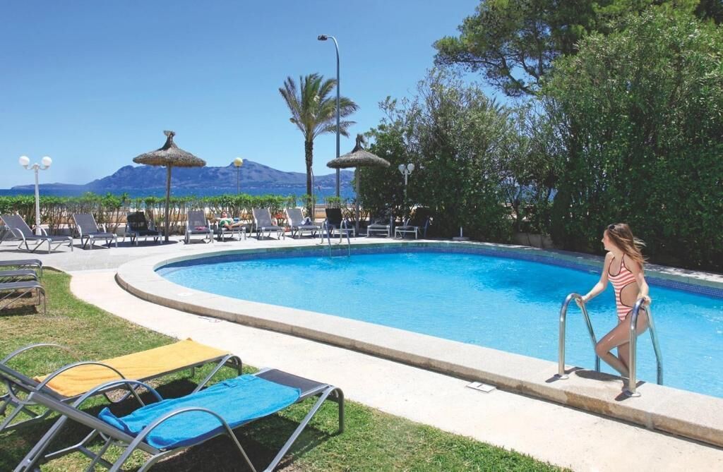 A swimming pool with lounge chairs and umbrellas, surrounded by greenery and a mountain in the background.