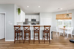 A modern kitchen with white cabinets, a central island with bar stools, a dining area with white chairs, and a window with blinds. - Spacious & Quiet Vacation Getaway, 15 Min to Beach (Wilmington)