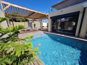 Ein Schwimmbad mit Holzterrasse und Liegestühlen, umgeben von viel Grün.