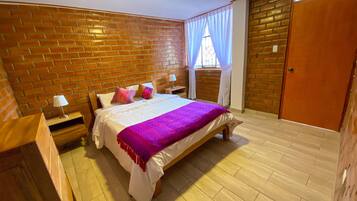 A bedroom with a brick wall, a bed with white bedding and a purple blanket, a nightstand with a lamp, and a wooden door.