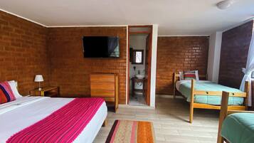 A hotel room with a brick wall, a bed with a red blanket, a bedside table with a lamp, a wooden dresser, a small bathroom, and a television mounted on the wall.