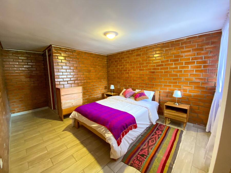 A bedroom with a brick wall, wooden floor, a bed with a purple blanket, two bedside tables with lamps, and a colorful rug.