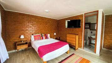 A bedroom with a brick wall, a bed with a white and pink bedspread, a wooden dresser, a TV mounted on the wall, and a bathroom visible through an open door.