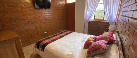 Un dormitorio con pared de ladrillo, un televisor de pantalla plana sobre la cama, una ventana con cortinas y un cabecero de madera.