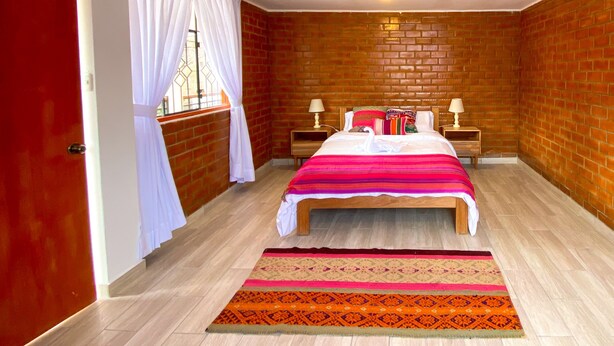 A bedroom with a brick wall, a bed with a striped bedspread, two bedside tables with lamps, and a window with white curtains. - Tupac Hostel Huaraz (Huaraz)