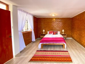 Une chambre avec un mur en briques, un lit recouvert d’une courtepointe à rayures, deux tables de chevet avec des lampes et une fenêtre avec des rideaux blancs.