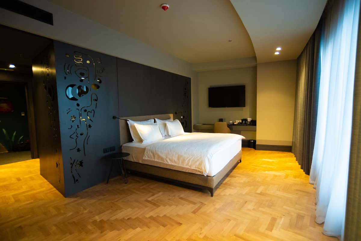 Une chambre d’hôtel moderne dotée d’un grand lit, d’un téléviseur à écran plat fixé au mur et d’un parquet en bois disposé en chevrons.