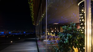 A rooftop restaurant with a view of the city at night, featuring a glass enclosure, tables set for dining, and a decorative plant. - Arka Art Hotel  (Farkë)