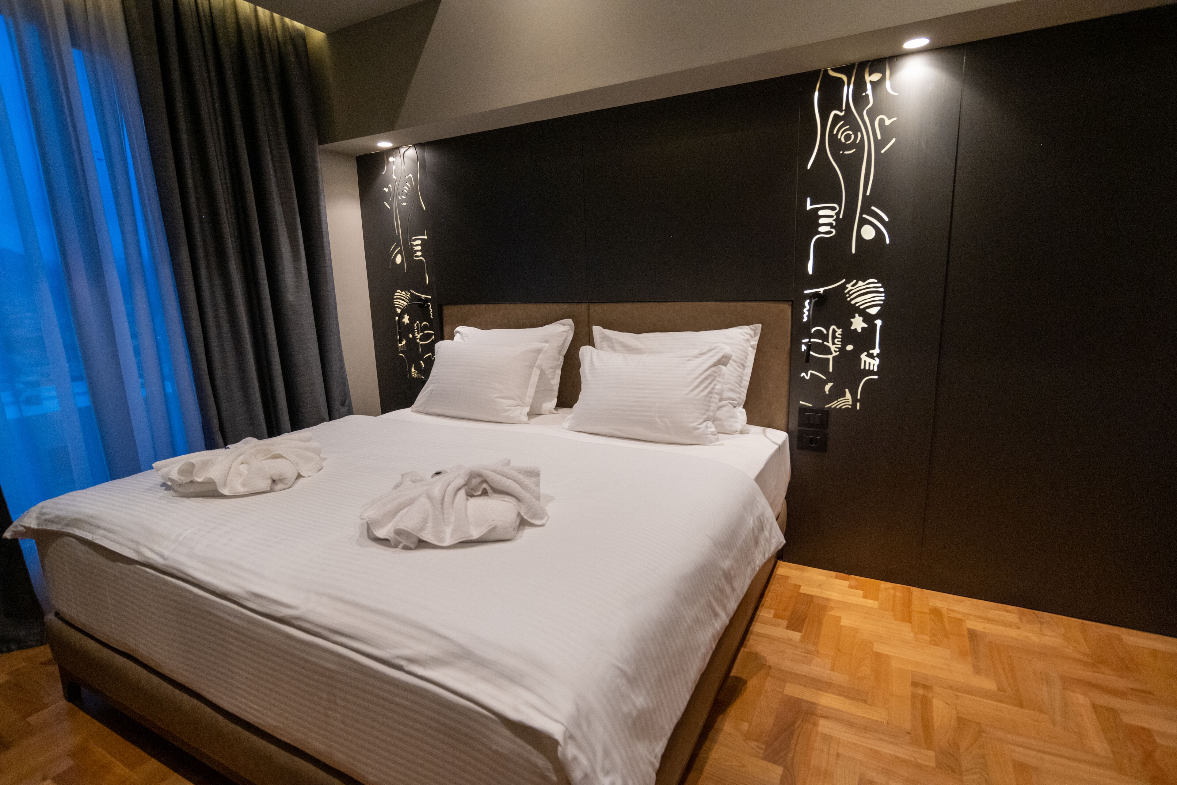 A hotel room with a large bed, wooden flooring, and a dark headboard with decorative wall art.