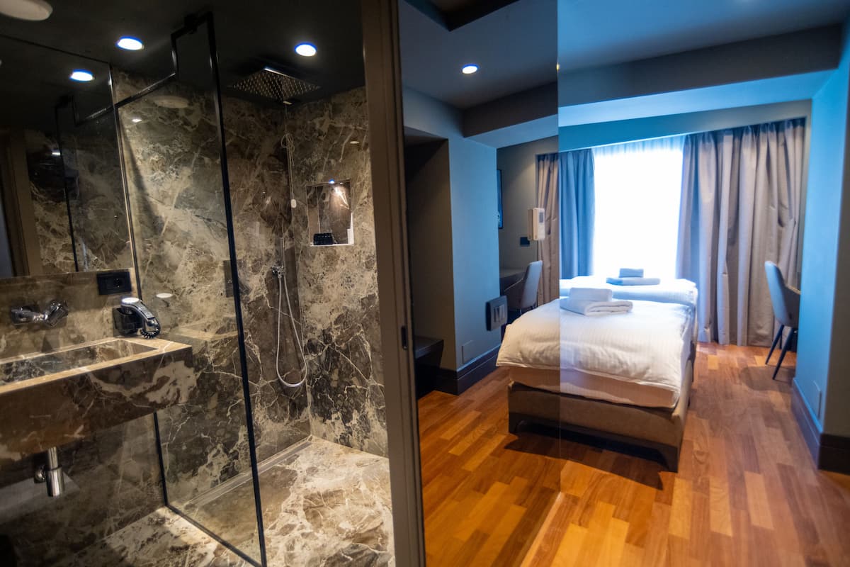 Une chambre d’hôtel moderne dotée d’une douche en verre, d’un plancher en bois, d’un lit recouvert d’une couette blanche et d’une fenêtre avec des rideaux.