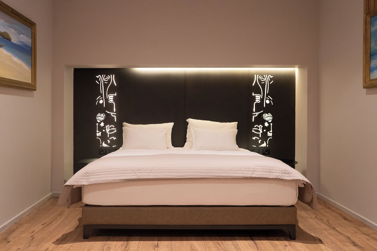 Une chambre à coucher moderne avec un grand lit, une tête de lit sombre ornée d’une calligraphie lumineuse et un plancher en bois.