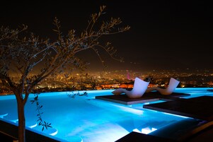 Una piscina infinita con vista a la ciudad por la noche, rodeada de sillones de descanso y un árbol.
