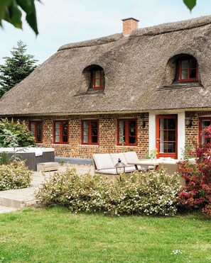A thatched-roof house with a brick exterior, a garden with flowering plants, and a seating area with cushions.