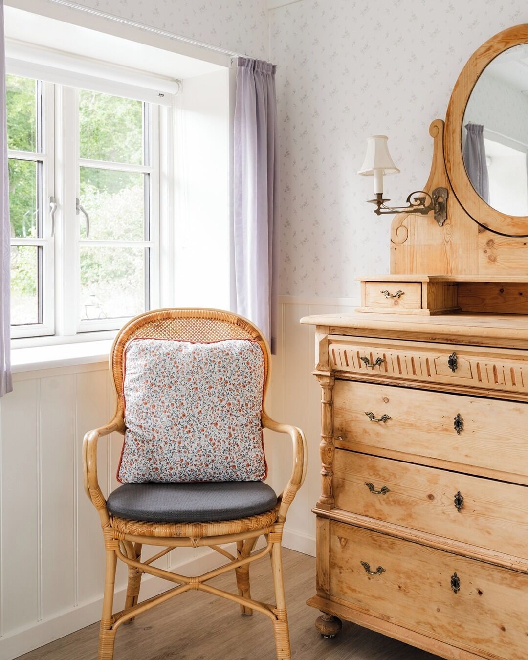 A room with a wicker chair, a wooden dresser with drawers, a mirror, and a window with curtains.