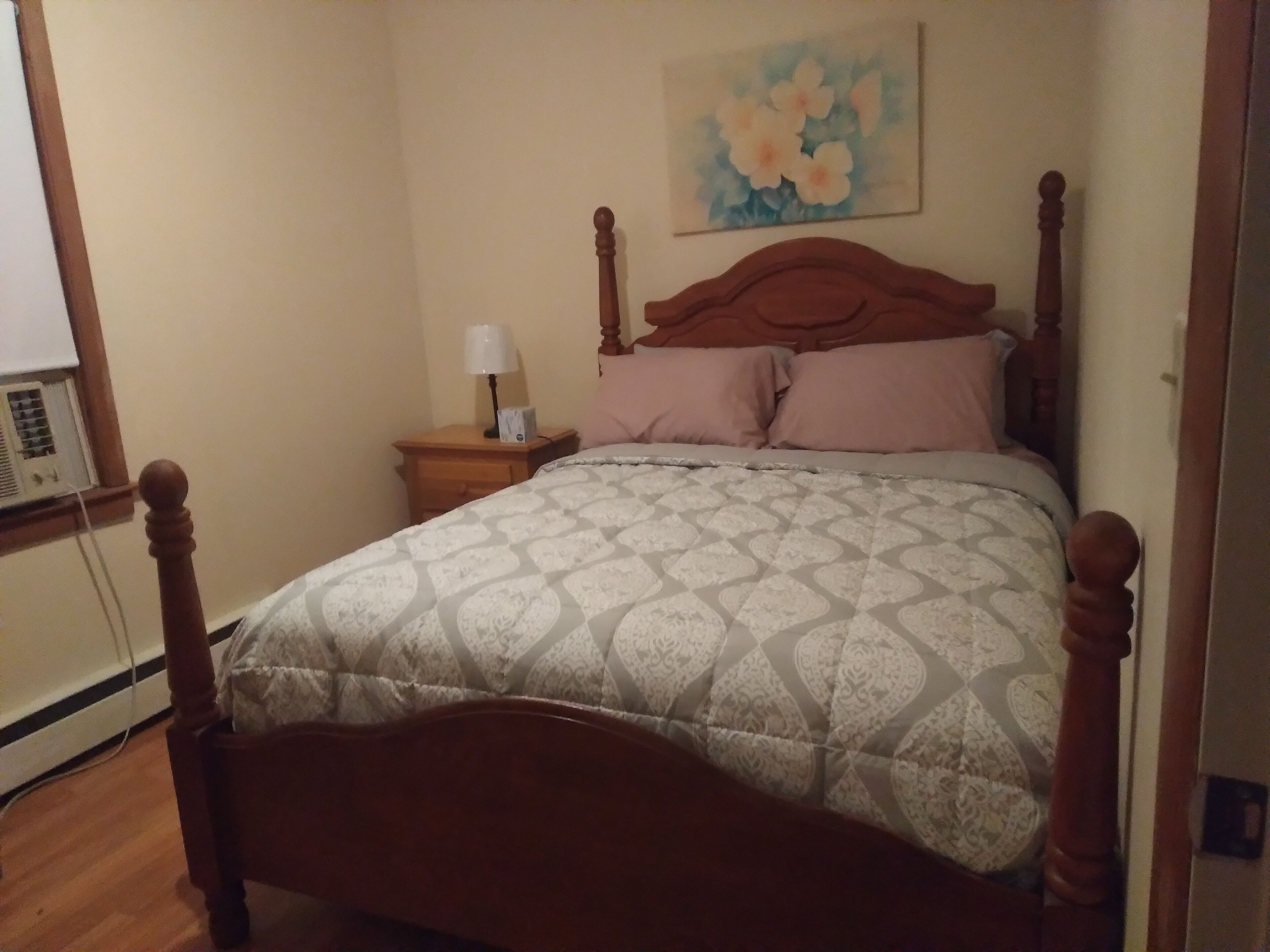 A bedroom with a wooden four-poster bed, a nightstand with a lamp, and a floral painting on the wall.