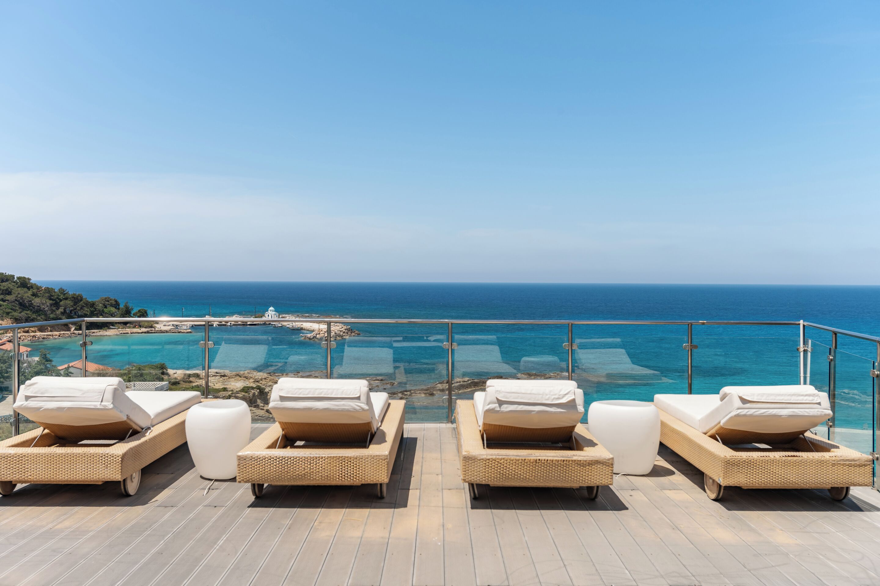 A terrace with four wicker lounge chairs and a glass balustrade overlooking a coastal area.