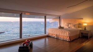 Un dormitorio con un ventanal grande con vistas al mar, una cama con almohadas, una mesita de noche con una lámpara y un pequeño banco con mancuernas.