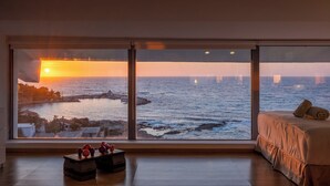 Una habitación con un ventanal grande que da a la vista de una puesta de sol sobre el mar, una cama y una mesita con objetos encima.