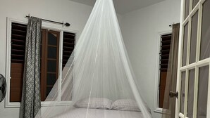 Een slaapkamer met een bed onder een witte hemel, beddengoed met een patroon, een klein tapijt en een plafondventilator.