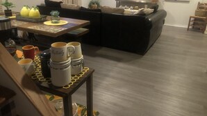 Una sala de estar con un sofá negro, una mesa de centro de madera y una zona de comedor con mantel estampado.
