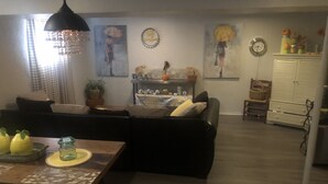 Una sala de estar con un sofá negro, una mesa de centro de madera y una mesa de comedor con adornos amarillos. Hay cuadros colgados en la pared y un reloj.