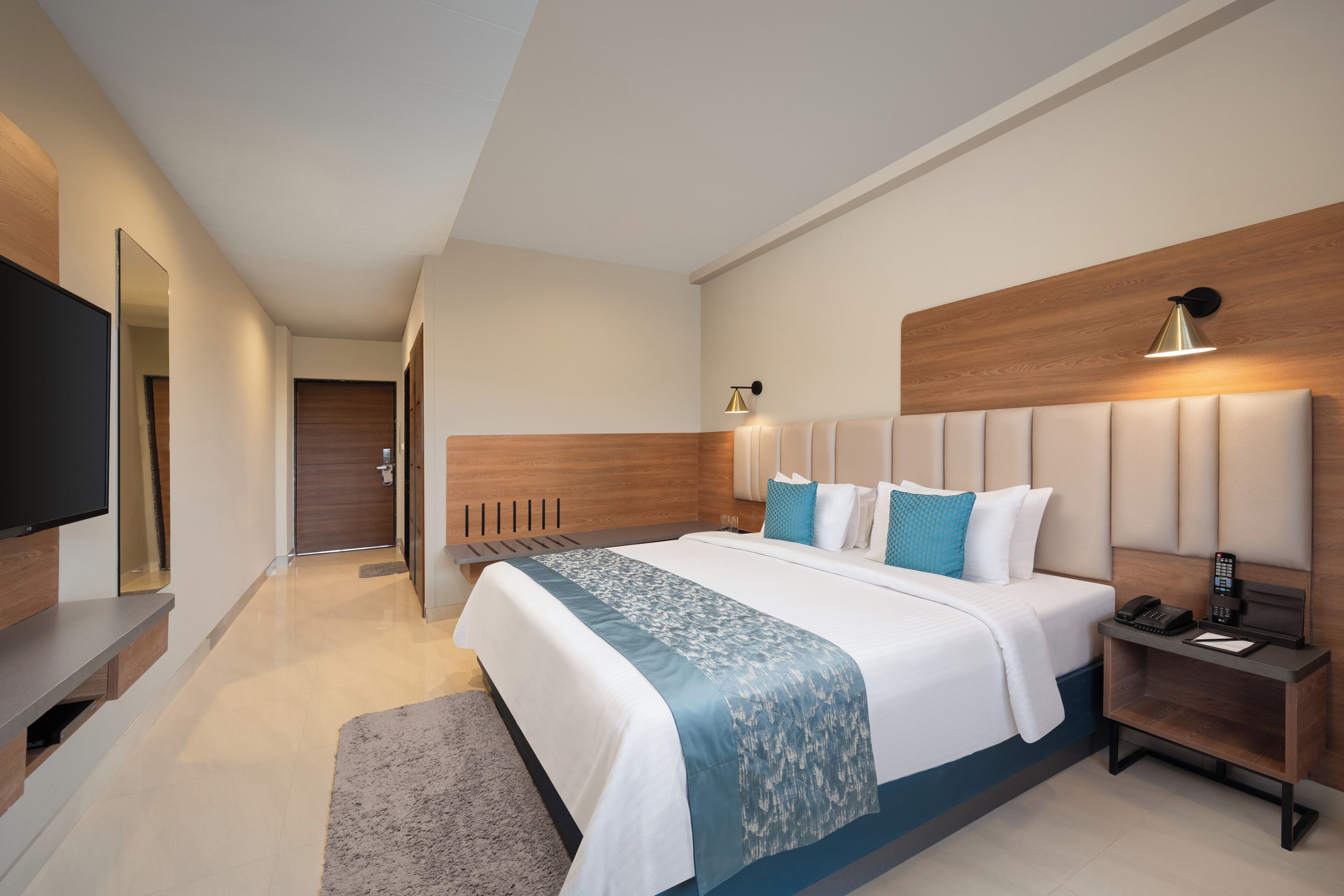 Ein modernes Hotelzimmer mit einem großen Bett, einem Nachttisch mit Lampe, einem Flachbildfernseher und einem hölzernen Kopfteil.