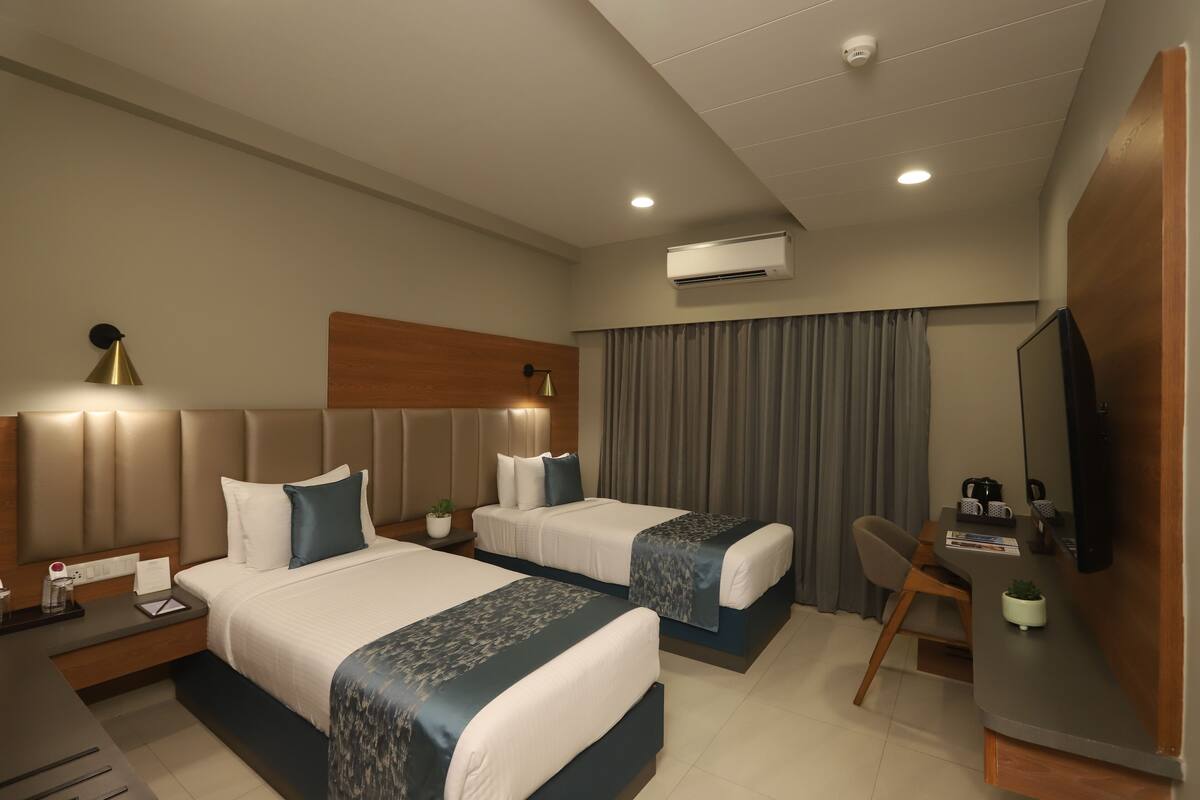 A hotel room with two beds, a desk, a chair, a TV, and an air conditioning unit.