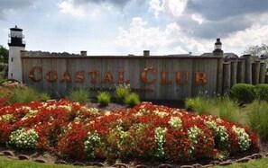 Et træskilt med 'COASTAL CLUB' skrevet på, omgivet af blomsterbede.