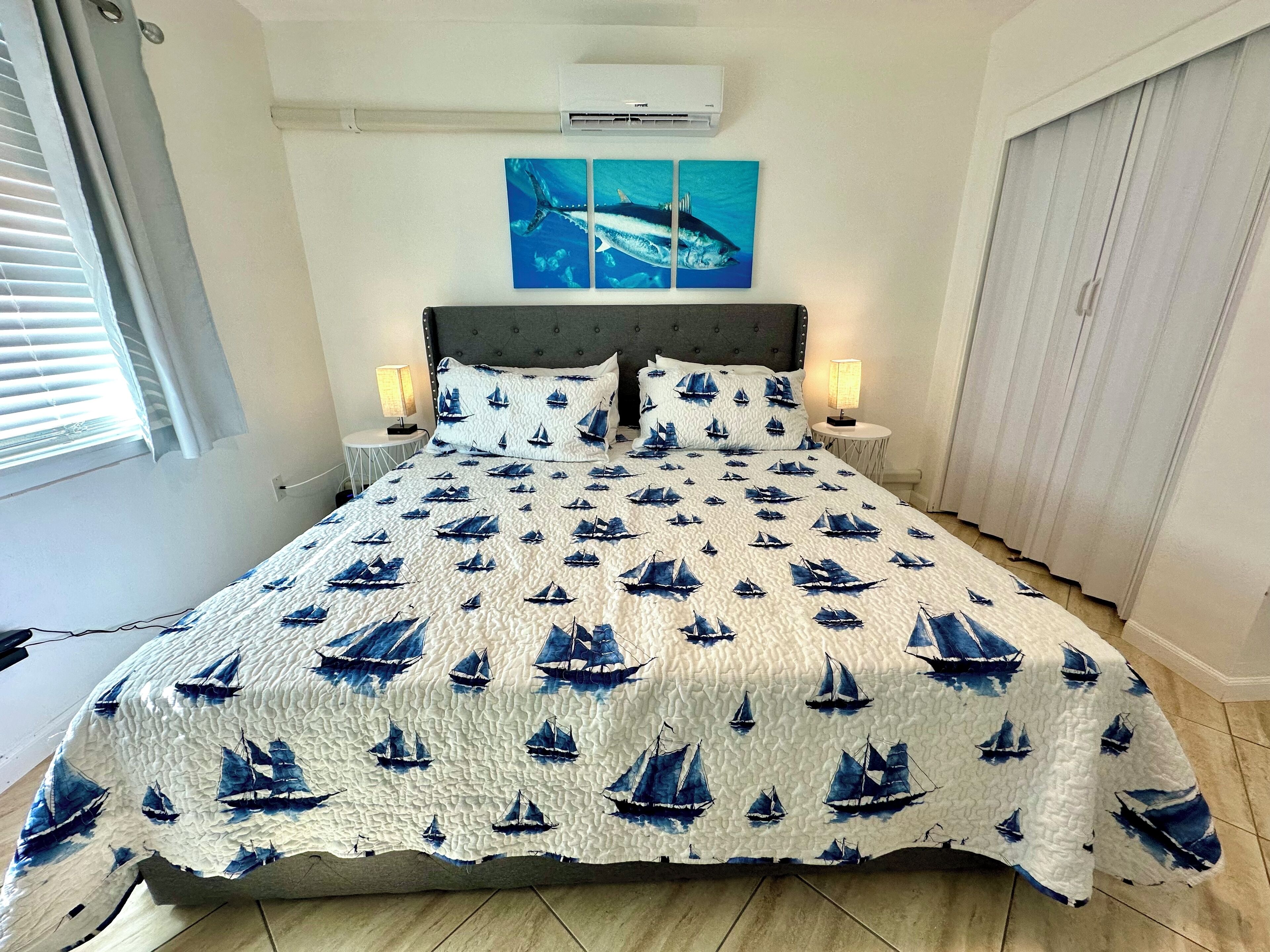 A bedroom with a bed, two bedside tables with lamps, a wall-mounted air conditioner, and a window with blinds.