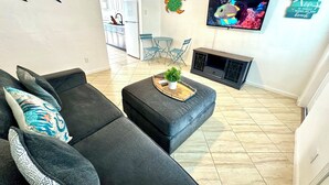 Una sala de estar con un sofá gris, una mesita con una planta, un televisor en la pared y una cocina al fondo.