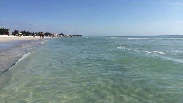 Una playa con agua clara y poco profunda, orilla de arena y algunas personas caminando.