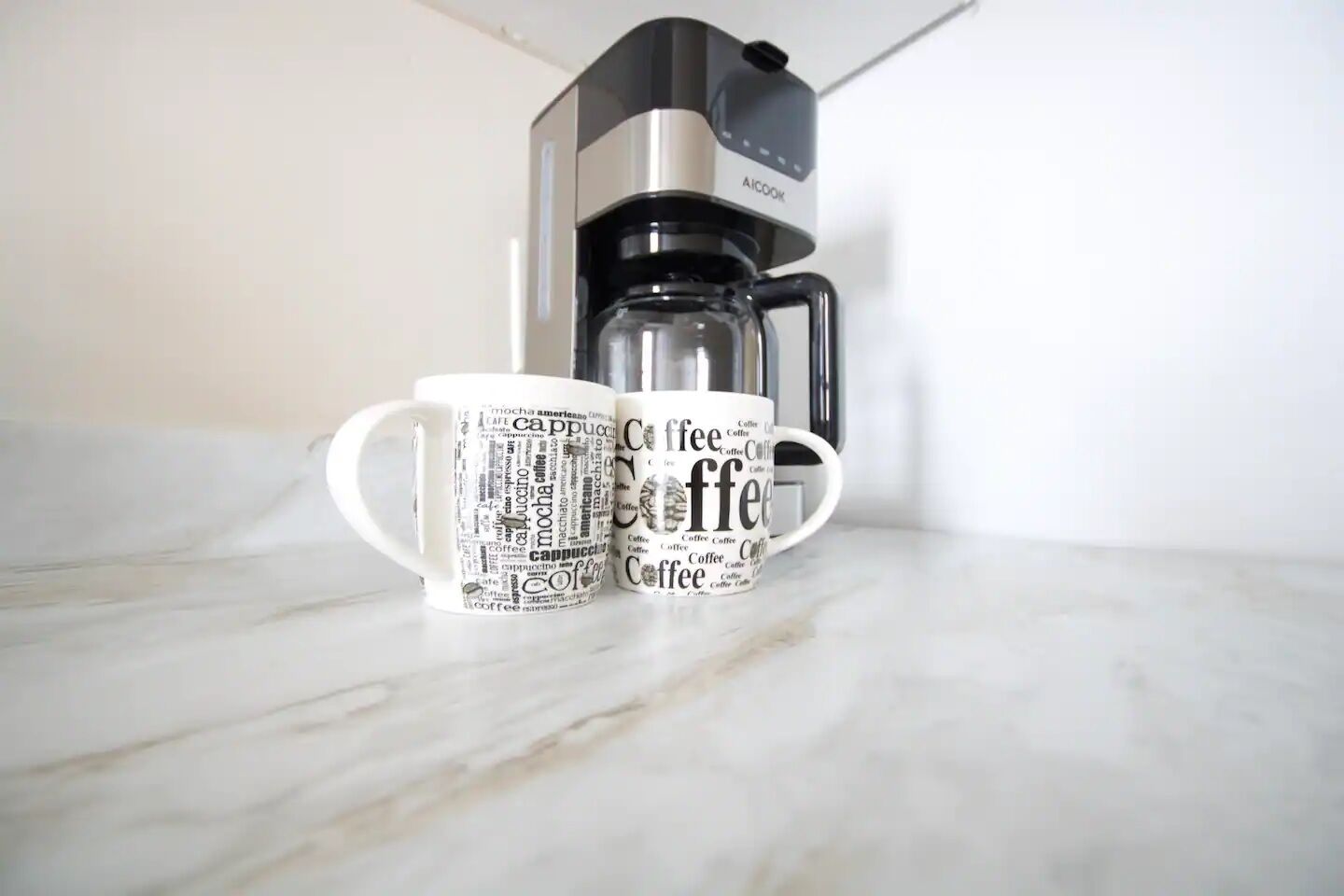Two coffee mugs with coffee-related text on a marble countertop, a coffee maker, and a white wall in the background.