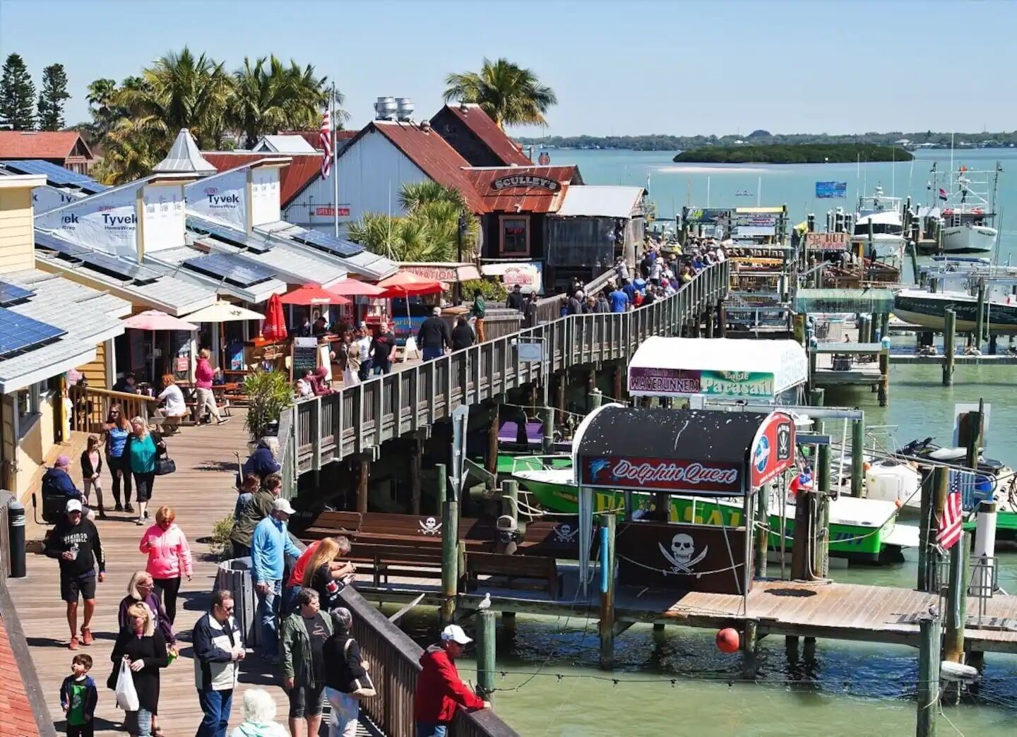 A bustling waterfront area with a wooden boardwalk, shops, and boats docked at the marina.