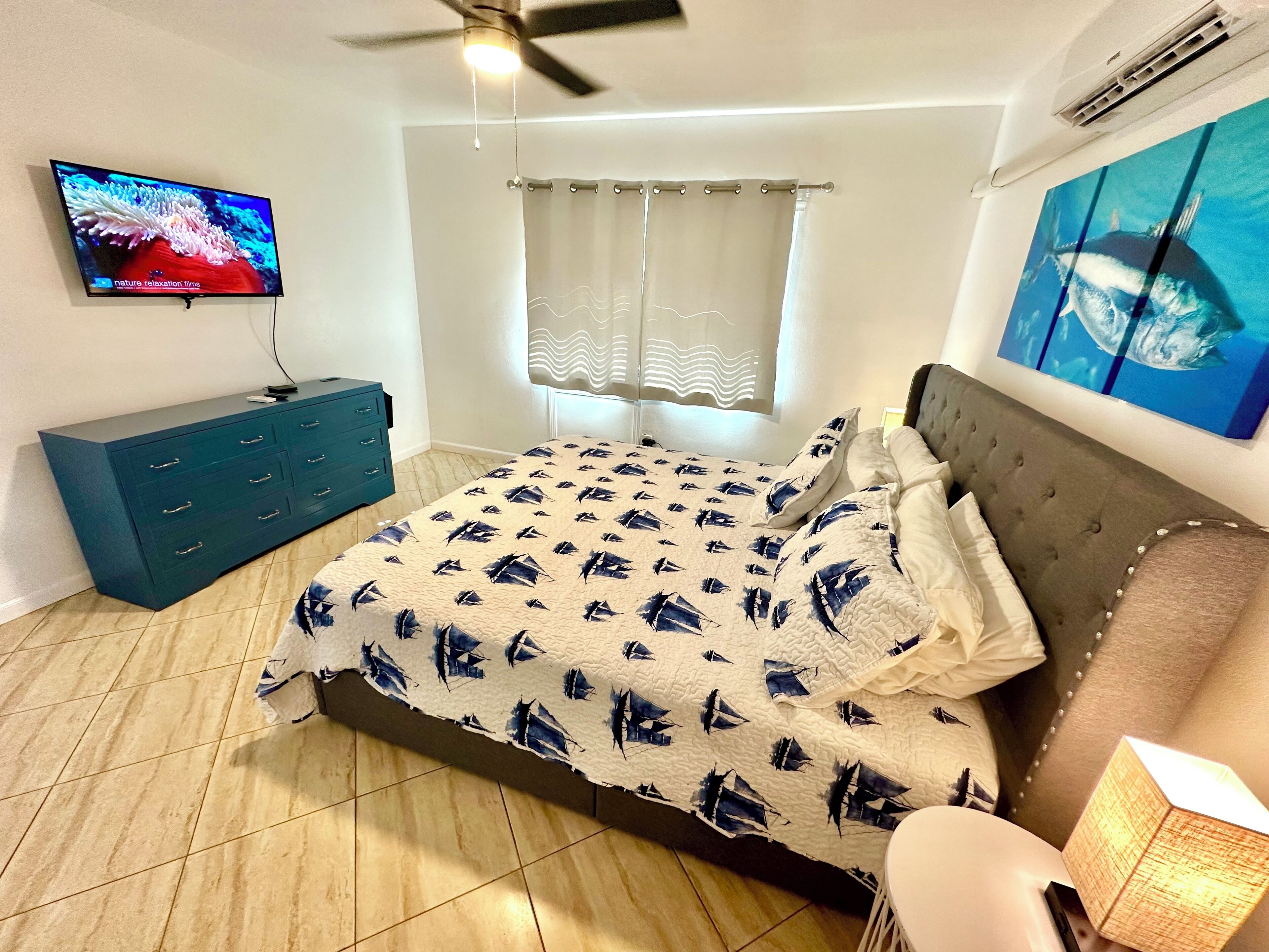 A bedroom with a bed, a television, a ceiling fan, a window with curtains, and a wall-mounted air conditioner.
